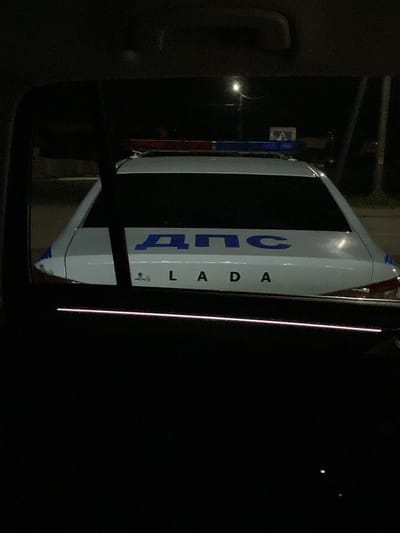 This is a vertical, night-time photograph taken in Kizner, Russia. The image focuses on the side of a white vehicle, possibly a van or a utility vehicle, which appears to be a LADA. The vehicle has a dark, rectangular section on its side with blue lettering. The visible text on the vehicle reads "ДПС" vertically, and below it, also vertically, "LADA". The "ДПС" lettering is large and blue. "LADA" is also in blue, with "L A D A" written vertically. There are small icons or symbols below the "LADA" text. The vehicle is parked outdoors in a dark environment, suggested by the lack of strong ambient light and the presence of artificial light sources. A street light is visible to the left of the vehicle, casting a soft glow. In the background, a pole with a sign that appears to have the letter "E" on it can be seen. The overall scene suggests a street or roadside setting at night. The dark background and the illuminated side of the vehicle create a contrast, highlighting the text and the vehicle itself. There are no people visible in the frame. The weather is not discernible from the image.
