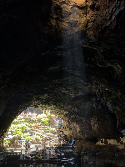 The image shows a view from inside a dark cave toward an illuminated, outdoor scene in Haría, Spain. The cave entrance frames the scene, casting much of the foreground into shadow. A beam of light cuts through the darkness within the cave. Outside, a large group of people are visible, some standing on what appears to be a complex of stone stairs and walkways. The architecture is surrounded by lush green vegetation, including plants and trees. Some reflections are visible on the right side.