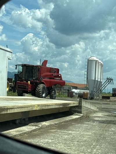 The image depicts agricultural equipment and structures, potentially a farm scene. A large red combine harvester is visible, along with a cylindrical grain silo supported by metal scaffolding. The background shows a cloudy sky. The scene appears to be next to a concrete structure. No people are visible. The precise location is specified as Straldzha, Bulgaria.