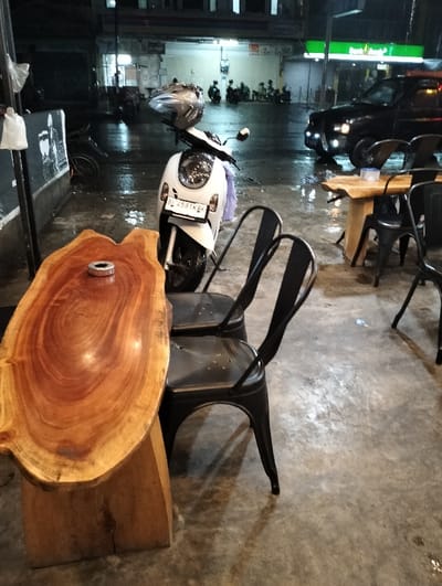 An outdoor cafe setting features a unique wooden table atop a stump, accompanied by two black metal chairs. A parked white scooter with a helmet on its seat is nearby. In the background, a street scene unfolds with people gathered and a black vehicle illuminated by headlights. The wet pavement reflects the surrounding lights, suggesting recent rain. A storefront with green signage is visible across the street.