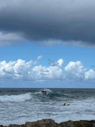 This image depicts a coastal scene in Le Moule, France, featuring two surfers under a dynamic sky. The upper third of the sky is dominated by a dense band of dark grey clouds, while the mid-section reveals bright blue sky interspersed with several distinct white cumulus clouds. The horizon line is visible separating the sky from the ocean.
The ocean exhibits dark blue-green waters with active wave conditions. A prominent breaking wave, characterized by white foam, extends across the mid-ground.
One surfer, clad in dark attire, is actively riding a white surfboard on the crest of a breaking wave, positioned slightly to the right of the image's center in the mid-ground. The surfer is leaning forward in a dynamic stance, generating white spray around the board. Further to the right and slightly behind the first surfer, a second individual is visible from the back, partially submerged in the water, positioned next to a light-colored surfboard. This second surfer appears to be stationary or paddling.
The immediate foreground consists of rugged, dark brown and grey rocks, indicating a rocky shoreline or coastal feature.