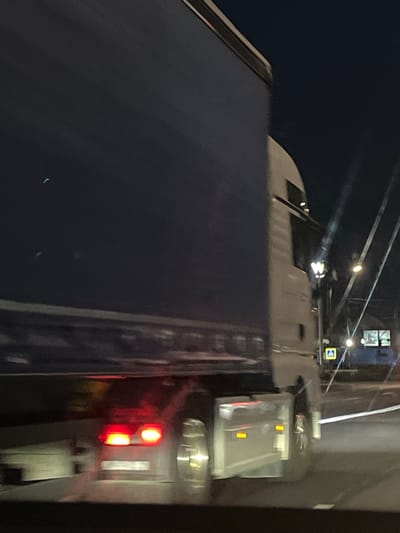 The image is a nighttime photograph taken from a vehicle in motion, likely on a road in Strumivka, Ukraine. The primary subject is a large, dark blue-sided truck, captured in profile and heavily blurred due to motion. The truck's cab is white and appears to be illuminated by streetlights and its own headlights. In the foreground, the truck's red taillights are visible and blurred. The background shows a dark sky with some faint lights, possibly streetlights or lights from buildings. There are also some blurred vertical lines that could be power lines or streetlights. A yellow pedestrian crossing sign with a black pedestrian symbol is visible on the right side of the image. The overall scene suggests a road at night, possibly in a semi-urban or suburban area.