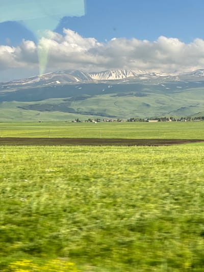 The image depicts a panoramic view of a vast, verdant landscape in the vicinity of Jamshlu, Armenia. In the foreground, lush green fields stretch across the frame, showing signs of agricultural activity with a dark strip of tilled soil running horizontally. The middle ground reveals rolling green hills dotted with a cluster of buildings, suggesting a small village or rural settlement. In the background, majestic snow-capped mountains form a dramatic backdrop under a bright blue sky adorned with fluffy white clouds. A peculiar light phenomenon, appearing as a green-hued, funnel-shaped cloud or light beam, emanates from the sky above the mountains. The scene conveys a sense of natural beauty and rural tranquility, possibly captured during a daytime journey.