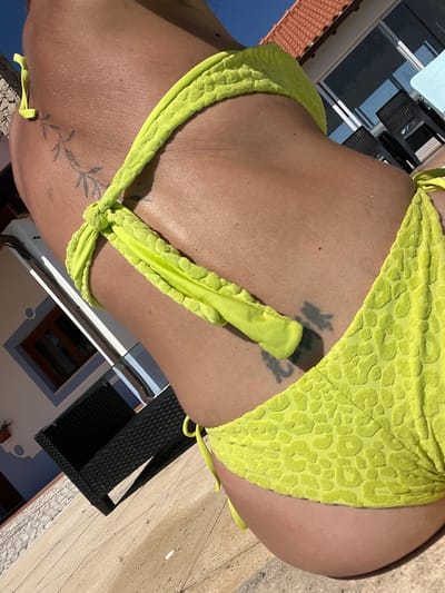 The image shows a close-up of a person's torso from the side, wearing a neon yellow bikini. The bikini has a textured pattern resembling leopard print. The person has at least two tattoos visible: one on the upper torso that appears to be a branch with leaves, and another on the lower torso that is darker and more abstract. The skin is tanned and appears to be in sunlight, with some light reflections and textures visible. The background is blurred but suggests an outdoor setting with a tiled floor, a building with a tiled roof, windows, and some outdoor furniture. The lighting indicates it is daytime with bright sun. There is no specific indication within the image that places it in Caldas da Rainha, Portugal.