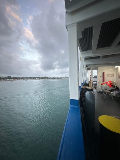 The image captures a view from the deck of a ferry at sea, looking towards a bay filled with sailboats and a distant shoreline with buildings and trees. The sky is overcast with dramatic clouds, some of which are tinged with pink and orange, suggesting either sunrise or sunset. The water is a choppy, deep teal color. On the right side of the frame, the ferry's deck is visible, with white support beams, a blue railing, and various objects including a yellow mooring bitt, life vests, tarps, and wooden benches. The overall scene conveys a sense of maritime transport and a coastal landscape. The location is identified as Pointe-à-Pitre, France, implying this is a ferry operating in or near that area.