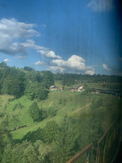 The image displays a rural landscape in Lazeshchyna, Ukraine, under a bright blue sky with scattered white cumulus clouds. The perspective is elevated, with a rusty metal railing partially visible in the lower right foreground. The immediate environment below consists of densely forested hillsides with various shades of green foliage. In the midground, rolling green hills feature patches of open grassland and interspersed trees. Multiple residential or farm buildings, characterized by light-colored walls and dark roofs, are visible on the hillsides, notably two clusters to the left and center-right. Fences are present along some of the grassy slopes. A dark-colored train or multiple carriages are visible moving horizontally on a railway track in the midground, extending towards the right. The background is dominated by distant, tree-covered hills. No people are present, and no specific actions or interactions are depicted.