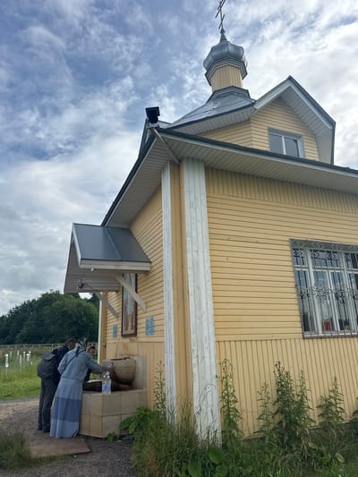 In the city of Рассвет, Russia, a yellow wooden building with a grey roof and a dome topped with a cross stands under a cloudy sky. The building has horizontal siding and a white vertical beam on its side. To the right of the image, a window with decorative iron bars is visible. In the foreground, a group of people are gathered at a stone fountain. One person, wearing a blue headscarf and a long blue skirt, is filling a plastic bottle with water from the fountain. Another person, with a grey backpack, stands beside them. The area around the fountain is overgrown with grass and weeds, and some white posts are visible in the background.