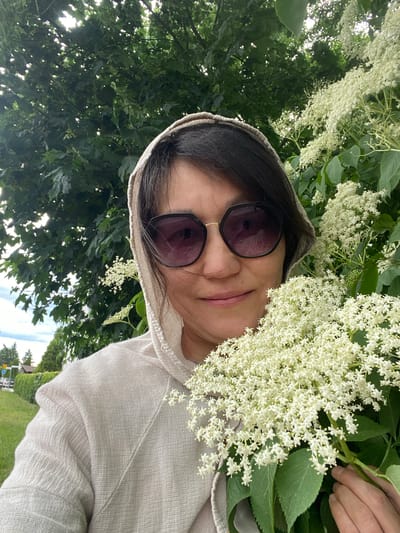 The image, captured in Dobieżyn, Poland, is oriented counter-clockwise and features a woman posing outdoors amidst vibrant green foliage and numerous clusters of small white flowers, likely elderflowers. She appears to be taking a selfie, holding a branch of these white blossoms close to her face.

The woman has dark hair and is wearing dark-lensed sunglasses and a light-colored, textured hooded top, possibly made of linen or cotton. A slight, pleasant smile is visible on her face. Her right hand is partially visible, holding the flower stem, and she wears a golden ring on her right ring finger.

The background is dominated by dense green tree leaves, with glimpses of bright sky indicating a clear, daytime setting. The overall scene conveys a fresh, natural environment, possibly a garden, park, or woodland area. The presence of abundant white flowers suggests spring or early summer. No visible text is present in the image.