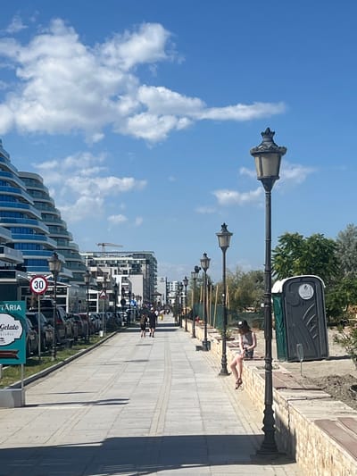 This media file shows a street scene in Năvodari, Romania. The image features a paved walkway lined with lampposts, a portable toilet, and modern buildings with a curved architectural style. Several people are visible, some walking and one lounging on a bench. Cars are parked along the street.
The time of day appears to be daytime, with clear blue skies and scattered clouds indicating fair weather. The presence of a portable toilet and people enjoying the outdoors suggests a public area, possibly a boardwalk or promenade.
Notable details include the distinctive architecture of the buildings, the vintage style of the lampposts, and a "30" speed limit sign. There is also text visible on signs, including "TARIA" and "Gelato Coffee," suggesting nearby businesses. The overall atmosphere seems relaxed and casual, with people enjoying a sunny day.
