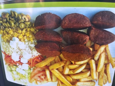 The image shows a plate of food, likely in Strombeek-Bever, Belgium. The plate contains falafel, french fries, a salad with shredded carrots and feta cheese, sliced tomatoes, corn, and sliced olives. The food is arranged on a white platter, with a green and black border at the top of the image. There are no people or animals visible in the image.