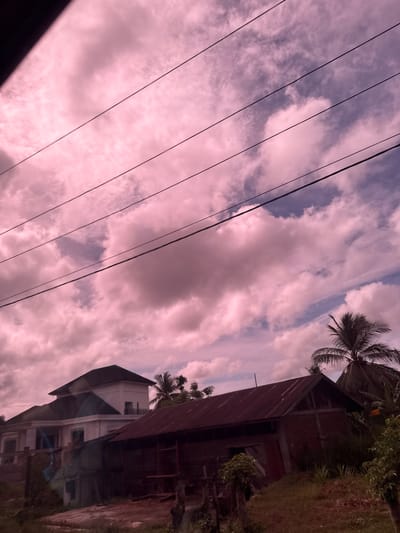 The image shows a cloudy sky with a pinkish hue, suggesting either a sunset or the effect of artificial lighting. Several power lines are visible cutting across the sky. In the foreground, there are two buildings. The larger building on the left is modern, with white walls and a dark roof, featuring columns and balconies. To its right, a smaller, older building with a rusted metal roof is visible. Palm trees are scattered in the background, and the overall environment appears to be a rural or semi-urban area. The location is identified as Buengbakjok, Indonesia. No people or specific actions are depicted in the image.