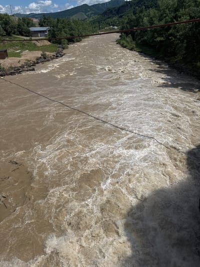 The image shows a wide, fast-flowing, muddy river in Каменномостский, Russia, under a bright, partly cloudy sky. The river dominates the frame, with turbulent water and white foam indicating a strong current. The riverbanks are lined with lush green trees and vegetation. On the left bank, there are some buildings and a small wooden structure. A red metal pipe, possibly part of a bridge or infrastructure, spans across the upper part of the image, with a thin wire or cable running across the river, casting a shadow on the water. The overall scene suggests a natural environment with some human presence, possibly during a period of high water levels.
