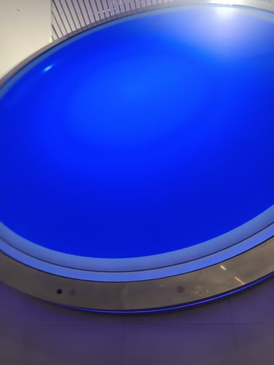 The image shows a large, circular blue display screen embedded in a wall, likely indoors. The screen is surrounded by a metallic frame with visible speakers or sensors. The wall beneath the screen is tiled. The location is Owle, India.