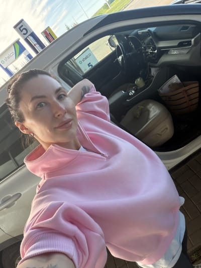 The image is a selfie taken by a woman inside a car, likely at a gas station in Ильинское, Russia. The woman, who appears to be young to middle-aged, is positioned diagonally across the frame, looking directly at the camera with a neutral to slightly smirking expression. She has light-colored eyes and her hair is styled in braids pulled back. She is wearing a pink full-zip hooded sweatshirt over a white undershirt, and a tattoo is visible on her left arm. Small hoop earrings adorn her ears.
The car's interior is partially visible, showing the dashboard, steering wheel, and part of the front passenger area. On the passenger seat or in the rear, a striped straw-like tote bag, a light-colored jerrycan or large plastic container, and a yellow wrapper are present.
Outside the car window, several gas station signs are visible under a bright, clear daytime sky. Prominently displayed are signs featuring the brand name "shelco" in green and blue logos, along with the number "5". Another yellow sign shows the text "АНЭКО" (ANEXO). The ground outside is paved with bricks. The scene suggests a casual stop, possibly for refueling, during clear weather.