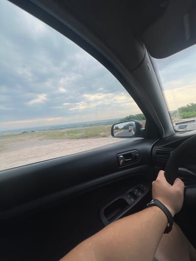 The image depicts a driver's perspective from inside a car, looking out the side window and at the steering wheel. The car is on a road with a slight incline, with a dry, dusty embankment on the left and sparse vegetation and trees on the right. In the distance, there appears to be a sprawling industrial area or town with smokestacks. The sky is overcast with dramatic clouds, suggesting a late afternoon or evening setting. A red car is visible in the side mirror, also on the road. The interior of the car is dark, with the driver's arm on the steering wheel and a smartwatch visible. The location is identified as being on a road leading into or out of Brestnik, Bulgaria.