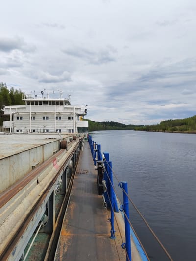 The image depicts a view from the deck of a river vessel, possibly a barge or a passenger ship, traveling along a waterway. The vessel's structure, including a white, multi-story cabin section with windows, is visible on the left side of the frame. A metal walkway with blue railings runs along the right side, adjacent to the water.
The body of water extends into the distance, bordered by forested areas on either side. The sky is overcast with grey clouds.
There is no verifiable indication of the location being Alekseevskoe, Russia, within the frame. No people are visible.