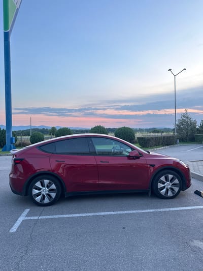 The scene depicts an outdoor parking area in Momkovo, Bulgaria, at either sunrise or sunset, indicated by the sky's blue, pink, and orange cloud formations. A deep red Tesla Model Y is parked, occupying a single asphalt parking space marked with white lines. The vehicle is presented in profile from its left side, featuring multi-spoke silver-toned wheels. A yellow and black wheel stop is positioned at the front right of the parking space. To the left of the parked car, a tall blue pole, partially supporting a green and white triangular sign and a blue box, ascends out of frame. To the right, a grey streetlamp with two light fixtures extends upwards. The background reveals a low, manicured hedge, an expansive agricultural field, and a distant mountain range under the colorful sky. No individuals are visible, and no actions are being performed.