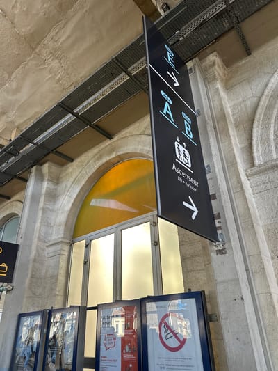This is a medium shot, taken from below, of a signpost in a public building, likely a train station. The sign is black with white and light blue text and symbols. It indicates directions to different "Voies" (platforms or tracks), labeled A and B. It also points to an "Ascenseur" (lift/elevator) with a wheelchair-accessible symbol. The sign is mounted on a metal bracket, and the building's architecture features rough-hewn stone and curved arches.

To the right of the signpost, there are framed notices. One prominently displays a red circle with a line through a cigarette, indicating "INTERDICTION DE FUMER" (No Smoking). Below this, there's text about fines and a helpline, "ALERTE SNCF" (SNCF Alert - a French railway company). Other framed notices show reflections of the surroundings, suggesting a bright, possibly sunny day, given the glare. The overall atmosphere is functional and informative, typical of a transportation hub. The time of day appears to be daytime, as indicated by the bright lighting and the general visibility. The setting appears to be indoors, under a covered walkway or terminal, with natural light entering from large windows or skylights.