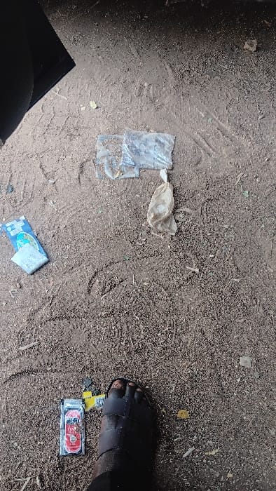 The scene depicts a reddish-brown dirt and gravel ground, presumed to be in Langtang, Nigeria, from an overhead perspective. A dark-skinned human left foot, encased in a dark, multi-strapped open-toed sandal, is positioned in the lower right quadrant. Various discarded items are scattered across the ground. In the lower left, adjacent to the foot, lies a silver and red rectangular packet, possibly for a consumable product, next to a crumpled yellow wrapper. In the upper-middle area, two pieces of clear, crumpled plastic sheeting are visible, along with a small, crumpled light-brown bag. To the left of this grouping, a blue and white sachet or package is present. The ground itself shows indistinct scuff marks and scattered small debris, including leaves. A dark, undefined object or shadow partially occupies the upper left corner of the frame.