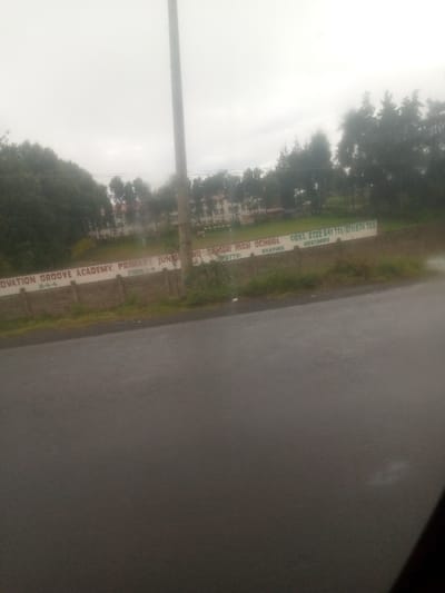 The image displays an overcast daytime scene of the Innovation Groove Academy, located in Sigona ward, Kenya, viewed from a slightly elevated position across a wet asphalt road. The foreground consists of a dark, reflective road surface, indicating recent precipitation. Beyond the road, a light-colored, low retaining wall separates the road from the school grounds. Painted prominently on this wall, in green and white, are the words "INOVATION GROOVE ACADEMY: PRIMARY JUNIOR AND SENIOR HIGH SCHOOL," "FORM 1-4," "8-4-4," "MOTTO: SHAPING DESTINES," and contact numbers "CONT: 0722 641 711 / 0711 874 165." Behind the wall, a green playing field with a white goalpost is visible. In the mid-ground, a tall, slender utility pole stands upright, extending towards the sky. The background features several multi-story school buildings with light-colored facades and darker roofs, partially obscured by dense green trees and foliage. The sky is uniformly light grey and overcast, with no direct sunlight visible. No individuals, specific actions, or interactions are discernible within the frame.