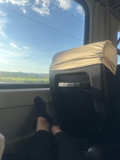 The image shows a view from inside a vehicle, possibly a train or bus, looking out the window. A person's legs, wearing black socks and pants, are visible in the foreground, resting on a surface. The window offers a view of a landscape with green fields, hills in the distance, and a bright blue sky with scattered clouds. The sun is shining, casting a warm light on the scene. A headrest, covered in a beige fabric, is prominent in the middle ground. The interior of the vehicle appears functional and utilitarian, with dark plastic elements and curtains visible. The context of Trastikovo, Bulgaria, suggests a rural or semi-rural setting outside a populated area.