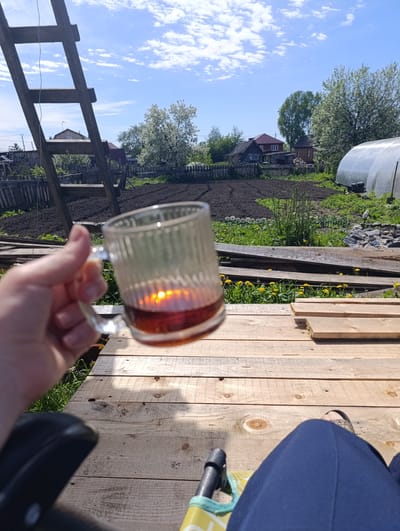 A first-person perspective photograph captures a tranquil outdoor scene in Yaya, Russia, on a bright, sunny day. The primary subject is a hand holding a clear, ribbed glass mug filled with dark liquid, likely tea, which brilliantly reflects the sun. The person is seated, with their legs, clad in dark blue trousers, and a shoe partially visible, resting on a wooden deck or platform made of light-colored planks. To the immediate left, the black armrest of a chair or similar seating is partially visible.

The background reveals a rustic garden setting under a clear blue sky dotted with scattered white clouds. A freshly tilled garden plot dominates the midground, bordered by a wooden fence. Beyond the fence, several rural houses with red and brown roofs are nestled amongst lush green and blooming trees, indicative of spring or early summer. A prominent wooden ladder leans upwards on the left side of the frame, while a large, tunnel-shaped greenhouse stands on the right. Patches of yellow dandelions are scattered across the grassy areas, adding a touch of vibrant color. The scene suggests a moment of peaceful relaxation in a rural or suburban environment. There is no visible text in the image.