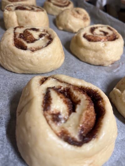 Ten unbaked cinnamon rolls are visible on a light grey parchment paper-lined baking sheet. Each roll consists of raw, light beige dough wrapped into a spiral, revealing a dark brown, granular cinnamon and sugar filling within its concentric layers. One roll dominates the immediate foreground, slightly out of focus, showcasing its intricate internal structure. The remaining nine rolls are positioned across the mid-ground and background, varying in sharpness. The background is indistinct, suggesting an indoor domestic setting. No individuals, actions, or interactions are discernible. The scene is located in a kitchen within a residence in Schmittweiler, Germany.