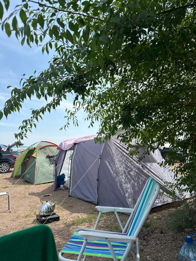 The image depicts an outdoor campsite situated near a body of water under a clear blue sky. The foreground features a colorful striped camping chair and a green blanket draped over another chair. Further back, a metallic kettle sits on a portable camping stove. Several tents are pitched on the sandy ground, including a large grey tent and a smaller green tent with a red trim. Behind the tents, the rear of a black car with its trunk open is visible. The campsite is partially shaded by the leaves of a tree, whose branches extend into the top of the frame. The setting appears to be a tranquil location, possibly a park or recreational area, in or near Kolodezny, Russia. There are no people visible in the image.