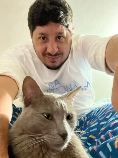 A selfie-style photograph from a high angle shows a man and a gray cat. The man is wearing a white shirt with writing in blue on it. The cat is gray and looking up at the camera. The background shows a surface or fabric with a blue pattern and white and orange dashes. The setting is unclear, there is no evidence to identify the location as Bernal Este, Argentina.