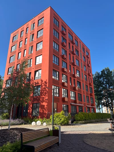 The media file depicts a modern, tall, salmon-colored residential building with numerous windows and square air conditioning units protruding from its facade. The building's architectural style is geometric and repetitive. To the right of the building, there is a paved walkway bordered by gravel and lush green landscaping, including shrubs, trees, and flowering plants. A wooden bench is visible in the foreground, adjacent to the walkway. The scene is illuminated by bright sunlight, suggesting it is daytime. The sky is a clear, vibrant blue, indicating good weather. The overall impression is one of contemporary urban living with integrated green spaces. The context provided places this scene in Kaluga, Russia. There is no visible text in the image.