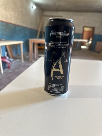 A close-up, eye-level, medium shot features a can of "Adrenaline Rush" energy drink positioned on a white table. The can is predominantly black with silver and gold accents. The text on the can indicates it is an energy drink with a "special complex of ingredients" for energy. A prominent "A" logo is visible, along with nutritional information. The background is blurred and appears to be an indoor construction site, with exposed walls, wooden structures, and doorways. The lighting is artificial, creating some shadows. The image is sharp, with focus primarily on the energy drink can. There are no people visible in the frame.