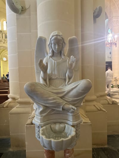 The image displays a white marble statue of an angel situated within a church in Għarb, Malta. The statue, centrally positioned and in sharp focus, depicts an angel seated cross-legged, hands raised with palms facing forward in a gesture of blessing or prayer. It wears long, flowing robes, has prominent wings, and a serene facial expression with a downward gaze. The angel rests upon a carved white marble pedestal which features an integrated scallop-shell-shaped font containing a clear glass vessel.

The background reveals a spacious church interior characterized by tall, light-colored stone or marble columns with decorative bases. The flooring consists of dark, possibly polished, tiles. To the left, rows of dark wooden pews extend into the mid-ground, where two individuals are partially visible, one wearing a striped garment, seated within them. An archway in the far left background leads to another section of the church, showcasing stained-glass windows and additional architectural elements. Modern white speakers are mounted on the columns overhead.

To the right of the statue, a person dressed in a full white suit is observed walking away from the camera, their back to the viewer, on the dark tiled floor. An ornate, multi-tiered metal chandelier with multiple light sources hangs from the ceiling in the upper right background. Another white wall-mounted speaker or fan is visible on a column in the mid-right. The scene is brightly illuminated, indicating ample light from both natural and artificial sources.