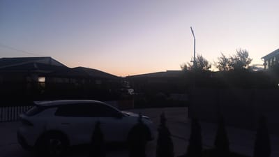 The image captures a suburban street scene at dusk in Choktal, Kyrgyzstan. A white SUV is parked in the foreground on a paved surface. In the background, several houses with dark roofs are visible, some with lights on inside. Trees and foliage are silhouetted against the light sky. A streetlamp stands tall to the right of the frame. The overall impression is one of quiet residential life as evening settles in.