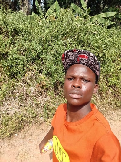 A young man poses for a selfie outdoors. He wears an orange t-shirt with yellow lettering and a skull-patterned flat-brimmed hat. He's holding a can in his left hand. Lush green vegetation forms a backdrop behind him, with banana trees visible in the distance. The ground appears to be a dirt path or road. Sunlight illuminates the scene, highlighting his face.