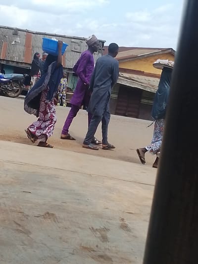 This media file captures a street scene in Ilero, Nigeria. Several people are walking across a paved road. On the left, a person wearing a dark headscarf and a patterned dress carries a blue plastic basin on their head. To their right, a man in a purple traditional outfit and a patterned cap walks alongside. Further right, another man in a grey traditional outfit is walking. In the background, several structures with corrugated metal roofs are visible, suggesting a marketplace or commercial area. A motorcycle is parked on the left side of the frame. The sky is overcast, indicating a cloudy day. The overall impression is of daily activity in a Nigerian town.