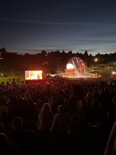 The image depicts an outdoor concert or performance taking place at dusk in Jonava, Lithuania. A large crowd of people is gathered, seated and standing, facing a stage. The stage is illuminated with stage lighting, and there are performers on it. A large screen is set up to the left of the stage, displaying a video feed of the performance. The sky is dark with a hint of twilight. The overall atmosphere is one of a lively public event.