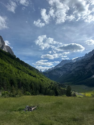 The image depicts a vast mountainous landscape under a partly cloudy sky in Shalë, Albania. In the foreground, a lush green meadow stretches out, dotted with tall grasses and a small pile of burnt wood and stones, suggesting a past campfire. To the left, a steep, heavily forested mountainside rises, densely packed with green trees. Further into the distance, a deep valley opens up, flanked by imposing, rugged mountains with rocky surfaces and patches of snow. The sky is a vibrant blue, punctuated by scattered white cumulus clouds, some of which are illuminated by sunlight. The overall scene conveys a sense of natural grandeur and tranquility.