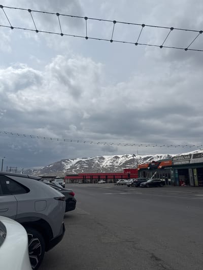 The photograph depicts a cloudy sky and mountainous terrain adjacent to a parking area and a roadside commercial building. The building has red vertical accents and features signage including "LPG" and "TIRES BATTERIES", with additional text in a non-English language. The parking area contains multiple vehicles, including a white SUV. A string of lights runs vertically from the top to the bottom of the image on the left. The image appears to be taken in Tsovagyugh, Armenia.