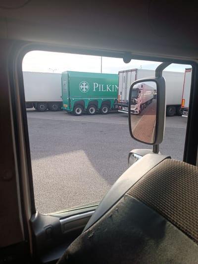 An exterior shot of a truck parking lot, possibly in Polesie, Poland. The image is taken from inside a vehicle, looking out a window and including a side-view mirror. In the foreground, the textured fabric of a seat and part of the truck's interior are visible. Outside, a line of semi-trailer trucks are parked on asphalt. One truck is predominantly green with the logo "PILKIN" prominently displayed. Other trucks are white or light-colored. The sky is overcast with diffused sunlight. The side-view mirror reflects another white semi-trailer truck and a brick-patterned walkway.