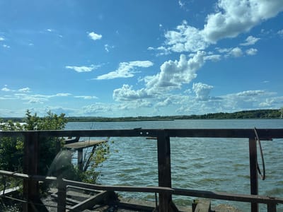 The image captures a view of a wide, choppy body of water under a bright blue sky with scattered white cumulus clouds. In the foreground, a weathered metal structure, possibly a railing or part of a dock, obscures the lower portion of the scene. The water stretches towards a distant tree line on the horizon, with hints of a flat landscape beyond. The scene appears to be a natural setting, possibly a lake or river. There are no people or animals visible in the image. The overall impression is one of a serene, perhaps slightly industrial, waterscape.