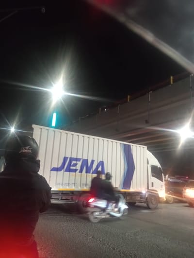 This media file depicts a nighttime scene in Kedungrejo, Indonesia, likely on a road beneath an overpass. The primary subjects include a large white truck with blue lettering that reads "JENA", partially obscuring a person in the foreground wearing a dark helmet and jacket. The truck has a license plate number visible, "082-200-757". In front of the truck, a motorcycle with two riders is in motion, captured with motion blur, suggesting movement. Further in the background, other vehicles are visible, also with some motion blur, indicating traffic. The lighting is characteristic of nighttime, with bright artificial lights from streetlamps or vehicle headlights causing lens flare effects. A bright blue light is visible above the truck, possibly a traffic signal or other form of illumination. The ground appears to be asphalt or gravel. The overall mood is one of a busy, active street scene at night.