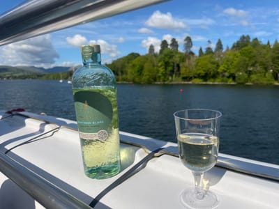 The image captures a serene moment on a boat, likely cruising on Lake Windermere in Bowness-on-Windermere, United Kingdom, as suggested by the lake, surrounding fells, and potential trees on an island visible in the background. A bottle of WISS WOLF Sauvignon Blanc, vintage 2023, stands next to a stemmed glass filled with the same wine. The scene suggests a relaxing afternoon or evening enjoying a glass of wine while surrounded by the natural beauty of the lake. The bottle's label indicates it is made using 100% consumer recycled glass. A metal railing suggests the boat is in motion, or docked. The sky is a mix of blue and white clouds.