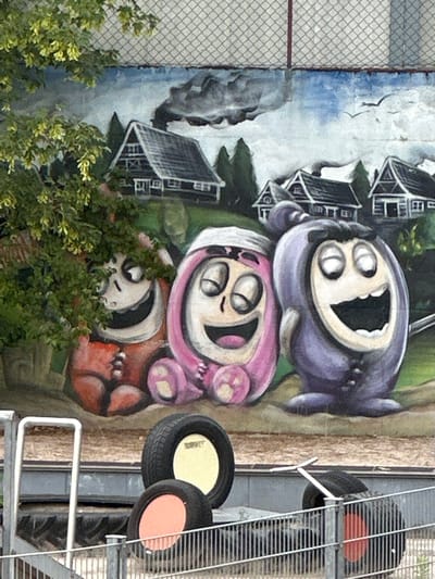 A vibrant outdoor mural dominates the center of the image, depicting three cartoon-like, rounded creatures stacked vertically. The top creature is purple, the middle one pink, and the bottom one, partially obscured by green foliage, is reddish-orange. All three share a similar design with wide-open mouths revealing teeth, and multiple large, white, circular eyes, conveying a joyful or surprised expression. The pink creature wears a white hood.

The mural's background features a serene village scene with several dark-roofed houses nestled among green trees under a cloudy sky, with hints of blue showing through and birds in flight.

To the left of the mural, a tall chain-link fence extends upwards, while on the right, various metallic structures are visible. These include a grey grated platform or staircase, a white handrail, and several black tires, some with yellow or orange centers, arranged near the metalwork.

Green, leafy bushes and trees are prominent in the foreground, especially on the lower left, partially covering the bottom portion of the mural.

The image appears to be taken during the daytime, likely under overcast conditions or late in the day. The setting suggests an urban or semi-urban environment, possibly a public wall in Herxheim, Germany, given the mix of street art, industrial elements, and natural foliage. There is no discernible text visible in the photograph.