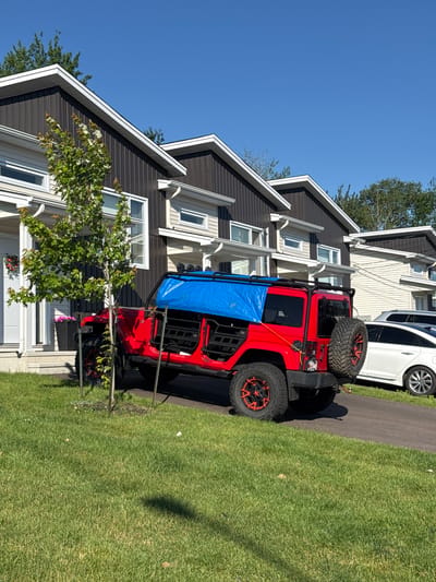 This image shows a red Jeep Wrangler parked on a residential street in Dieppe, Canada. The Jeep is covered with a blue tarp, which is secured by ropes and some kind of frame. The vehicle has large, off-road tires with red accents on the rims. In the background, there are several modern townhouses with dark brown siding and white trim. A small tree with green leaves is planted in front of the Jeep. To the right of the Jeep, a white car is parked in the driveway of one of the townhouses. The scene is bathed in bright sunlight, casting shadows on the green lawn. The sky is clear and blue.