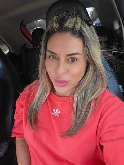 The image shows a woman in the driver's seat of a car. She is wearing a coral-colored sweatshirt with the Adidas logo on it. Her hair is blonde and pulled back. She is looking at the camera with her mouth slightly open. The car appears to be a compact model, with black leather seats and a red interior trim. The background is blurred, but it suggests an outdoor setting with trees and other vehicles visible. The image quality is sharp, with good lighting and detail. The car's interior suggests it is a modern vehicle. The woman's makeup is subtly applied, enhancing her features. The overall impression is a casual selfie taken during a drive. The specific location within Carpi, Italy, cannot be determined from the visual information provided in the image.