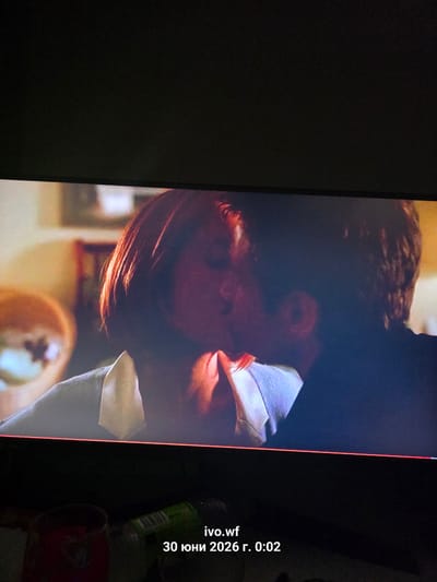 The image displays a screenshot of a video, showing a close-up of two people kissing. The lighting suggests an indoor setting, possibly a living room or bedroom. The focus is on the couple's faces and upper bodies, with a slightly blurred background. In the foreground, at the bottom of the frame, there are some objects on a dark surface, including a red glass and a green bottle with a label. Text overlay on the bottom right corner reads "ivo.wf 30 юни 2026 г. 0:02". There is no discernible information in the image to indicate the specific location within the city of Chal, Bulgaria.