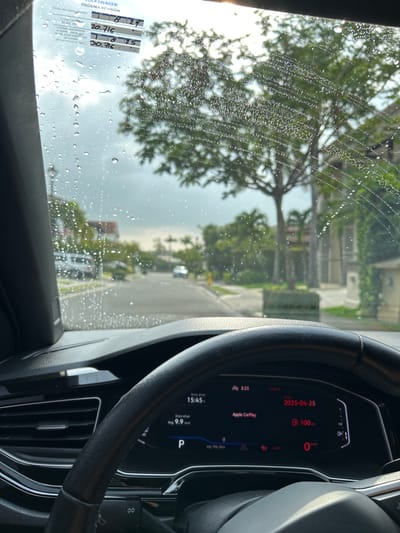The image shows a view from the driver's seat of a car. The car is parked on a street with houses on either side. The windshield is covered in raindrops, and the image is blurry due to the rain. The driver's side door is visible on the left side of the image, and the dashboard of the car is visible on the right side. The dashboard includes a digital display showing the time, temperature, and other information. The location is in Río Oro, Costa Rica. The coordinates are latitude: 9.945, longitude: -84.201.