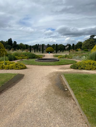 This is a landscape photograph taken from a slightly elevated perspective, looking down a gravel path that curves through a well-maintained garden. The image is oriented vertically, with the path dominating the foreground and middle ground. To the left of the path is a lush green lawn interspersed with flower beds filled with a variety of plants, including some taller, spiky shrubs and smaller, bushy plants. Further back, a formal garden layout is visible with manicured topiary trees and a distinctive circular water feature. In the distance, a large building with architectural features reminiscent of a stately home or castle can be seen, surrounded by more trees. The background is filled with a dramatic sky, characterized by a mix of dark, heavy clouds and patches of lighter sky, suggesting overcast or changing weather conditions.
The scene depicts a serene and picturesque garden setting, likely a public park or a private estate. The presence of the gravel path, manicured lawns, and formal planting suggests a carefully designed and tended landscape. The time of day appears to be daytime, given the natural light, but the cloudy sky might diffuse the sunlight. The location context of Stafford, United Kingdom, implies a temperate climate. There are no people or animals visible in the frame, and no text is present. The overall mood conveyed by the image is one of tranquility and natural beauty, with a touch of historical grandeur suggested by the distant building.