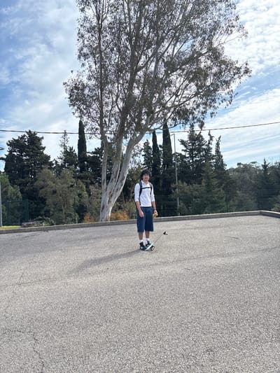 On a sunny day in Fréjus, France, a young man stands on an asphalt road next to a large tree with a skateboard at his feet. He is wearing a white long-sleeved shirt, dark blue shorts, and black and white sneakers. He has dark curly hair and is facing the camera with a neutral expression. He is holding a small yellow object in his right hand. A backpack strap is visible across his chest.

Behind him, there is a fence and a line of trees. The sky is partly cloudy. The road appears to be a public street or path in a park or residential area. The image is taken at a slightly low angle, and the perspective is distorted, as if taken with a wide-angle lens.
