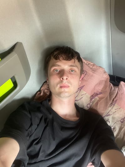 A man with light brown hair and green eyes is lying down and looking at the camera. He is wearing a black t-shirt. He is resting on a pink and beige striped pillow with a floral pattern. The background appears to be a light gray surface, possibly upholstery or fabric. In the foreground, there is a gray plastic object with a bright green insert. The lighting suggests it could be daytime, but there are no specific cues for time of day or weather. The image appears to be a selfie taken indoors. No text is visible.